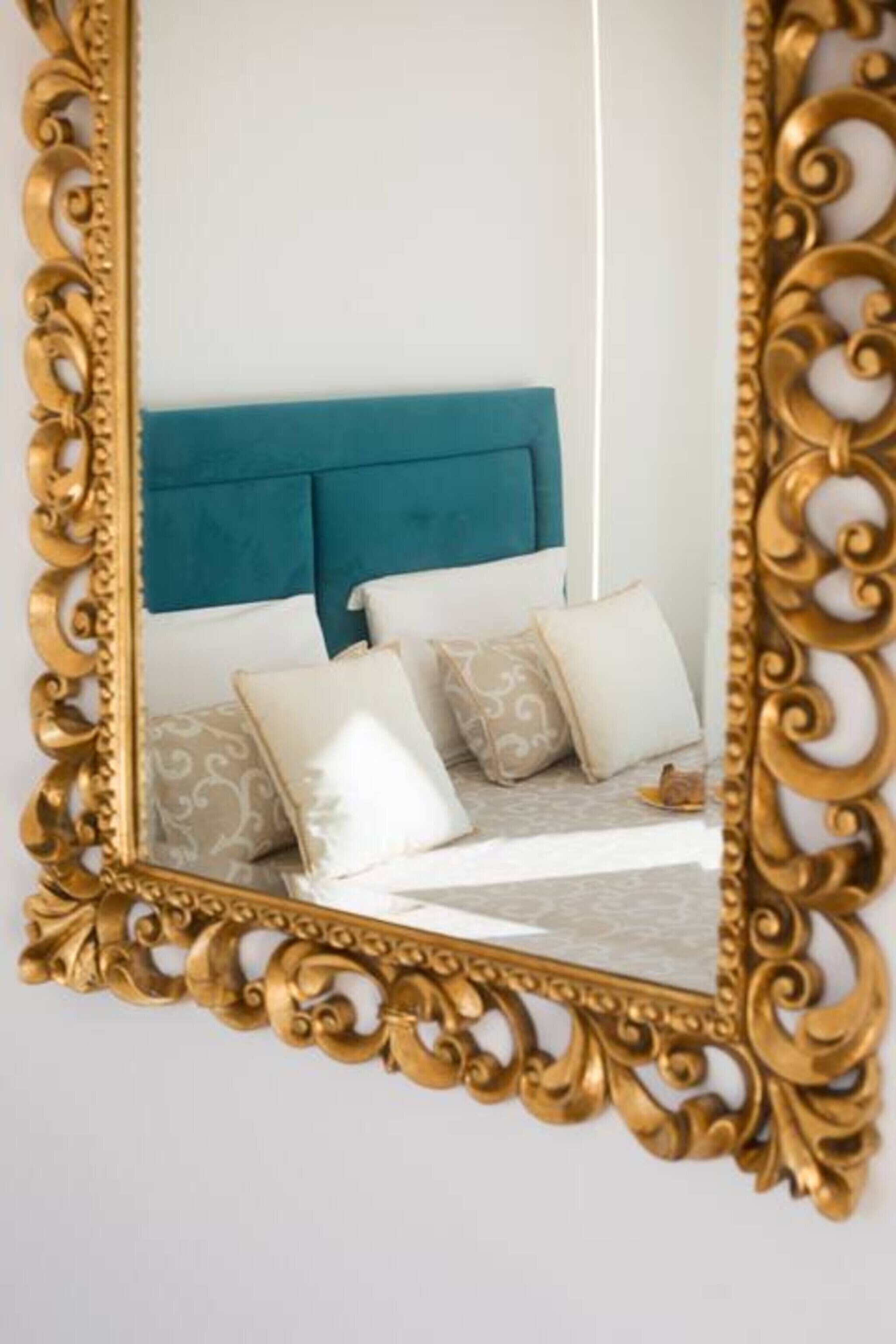 Uma cama com cabeceira verde-água, almofadas brancas e estampadas, e um espelho com moldura dourada e ornamentada.