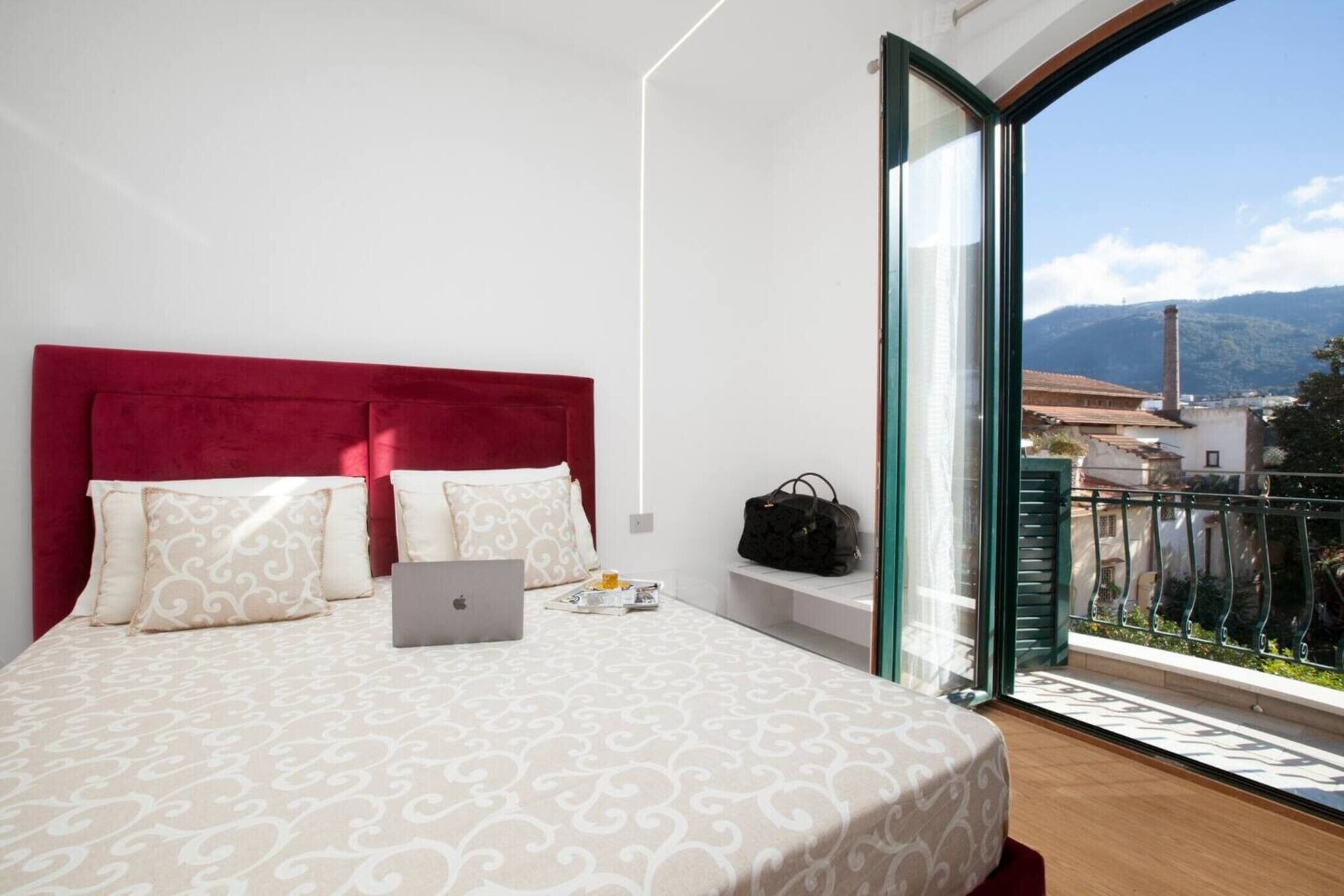 A bedroom with a red headboard, a laptop, a handbag, and a balcony with a view of buildings and mountains.