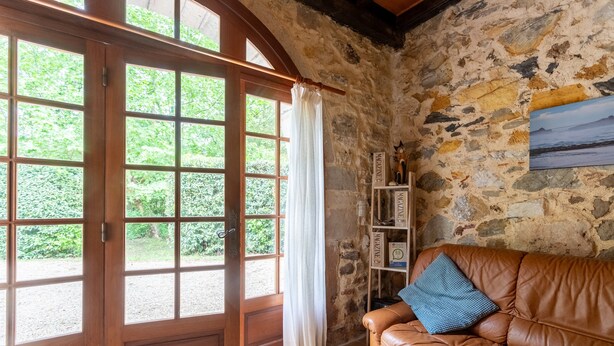Ein Innenraum mit einer Steinwand, einer Holztür, die in einen Garten führt, einem braunen Ledersofa mit Kissen und einem Regal mit verschiedenen Gegenständen.