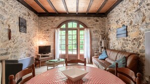 Ein Zimmer mit Steinwänden und Holzboden, einem Fernseher, einem Tisch mit kariertem Tuch, einem Ledersofa und einem großen Bogenfenster.