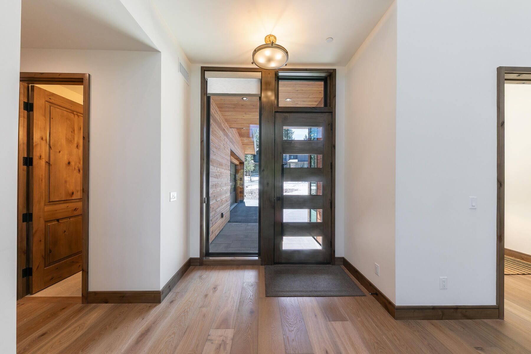 A hallway with wooden flooring, a wooden door on the left, and a glass door leading to a covered outdoor area.