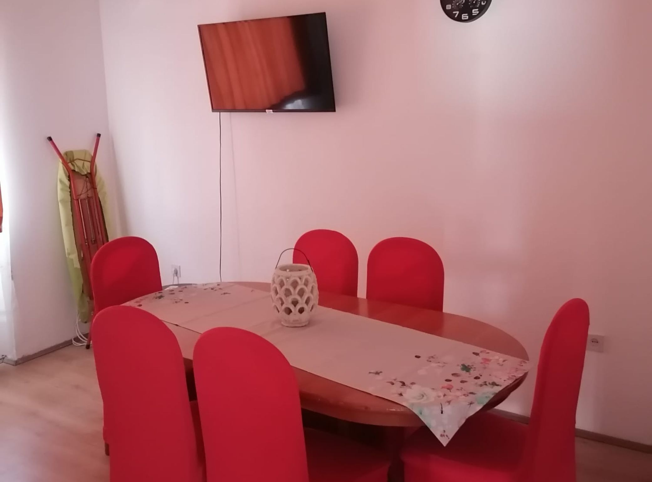 Ein Essbereich mit einem runden Tisch, sechs roten Stühlen, einer Vase und einem an der Wand befestigten Fernseher.