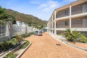 A multi-story residential building with balconies, a paved driveway, and a mountain backdrop.