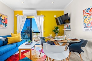 A modern living room with a blue sofa, a glass dining table, and a yellow accent wall.