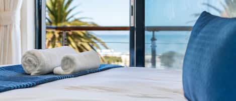 A neatly made bed with rolled white towels and a blue pillow, a blue striped blanket, and a view of palm trees and a clear sky through large windows.