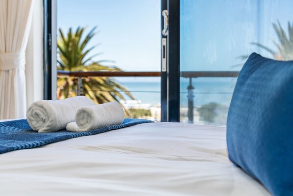 Una cama bien hecha con toallas blancas enrolladas, una almohada azul y una manta a rayas azules, con vistas a palmeras y un cielo despejado a través de amplias ventanas.