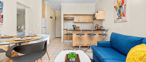 A modern living room with a blue sofa, a glass dining table with chairs, and a kitchen in the background.