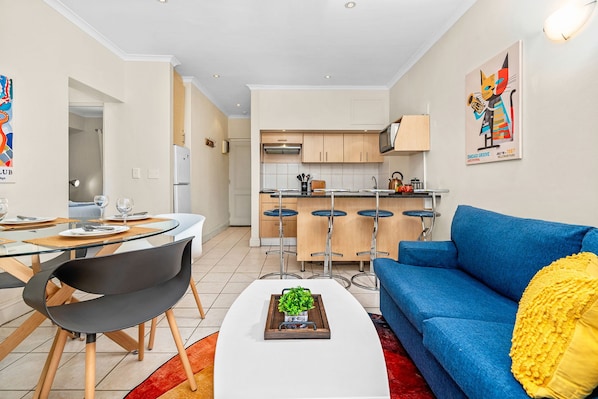 Una sala de estar moderna con un sofá azul, una mesa de comedor de vidrio con sillas, y una cocina al fondo.