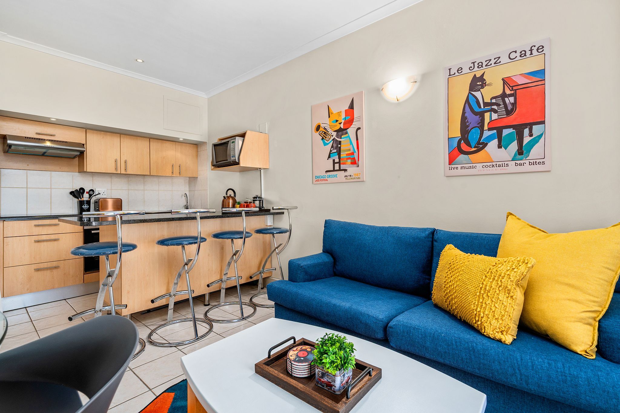 A modern kitchen with bar stools, a blue sofa with yellow cushions, and a coffee table with a tray.