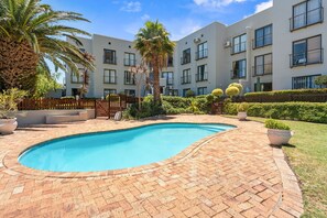 A swimming pool surrounded by a brick patio, with a multi-story building in the background. - Gordon's Village View (Cape Town)