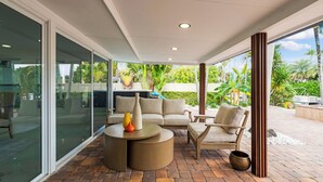 A modern outdoor patio with a sofa set, a round table, and a large glass door leading to a garden. - Waterfront 4BR Retreat w/HotTub Dock & Outdoor Bar (Fort Lauderdale)