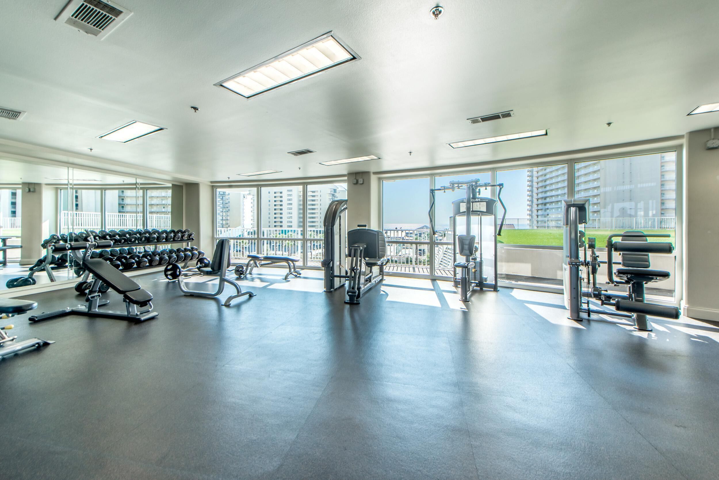 Ett rymligt gym med olika typer av träningsutrustning, stora fönster och utsikt över byggnader.