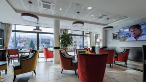 Un moderno vestíbulo de hotel con sillas rojas y amarillas, una gran pantalla de televisión y vistas de la ciudad a través de amplias ventanas.