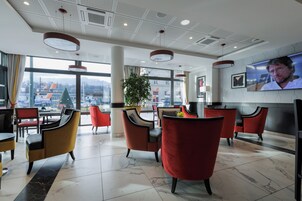 Um hall de hotel moderno com cadeiras vermelhas e amarelas, uma televisão grande e vista para a cidade através de amplas janelas.