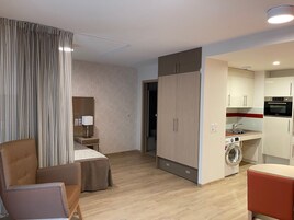 Quarto de hotel moderno com cama, escrivaninha, cadeira, armário, máquina de lavar e cozinha compacta.