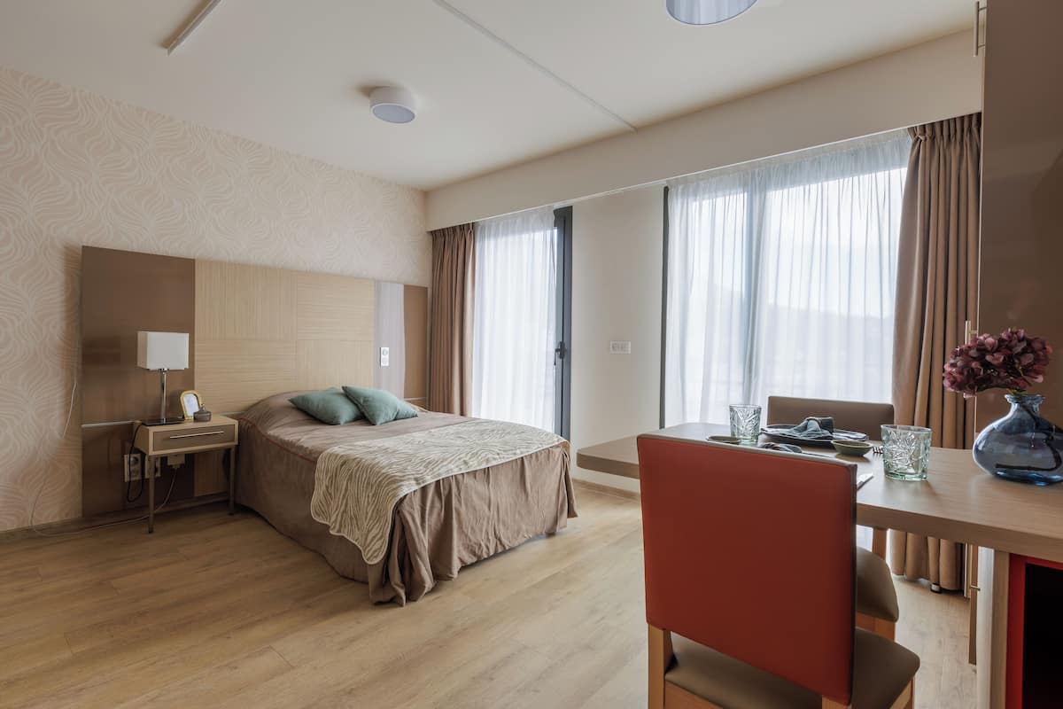 Una habitación de hotel con cama, mesita de noche, lámpara y un área de comedor con mesa y sillas.