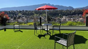 Una terraza con sombrilla roja, dos sillas y una mesa con vistas a un pueblo y montañas.