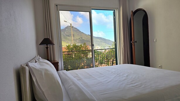 A hotel room with a bed, a lamp, a large mirror, and a balcony with a mountain view. - K&K Apartments (Machico)