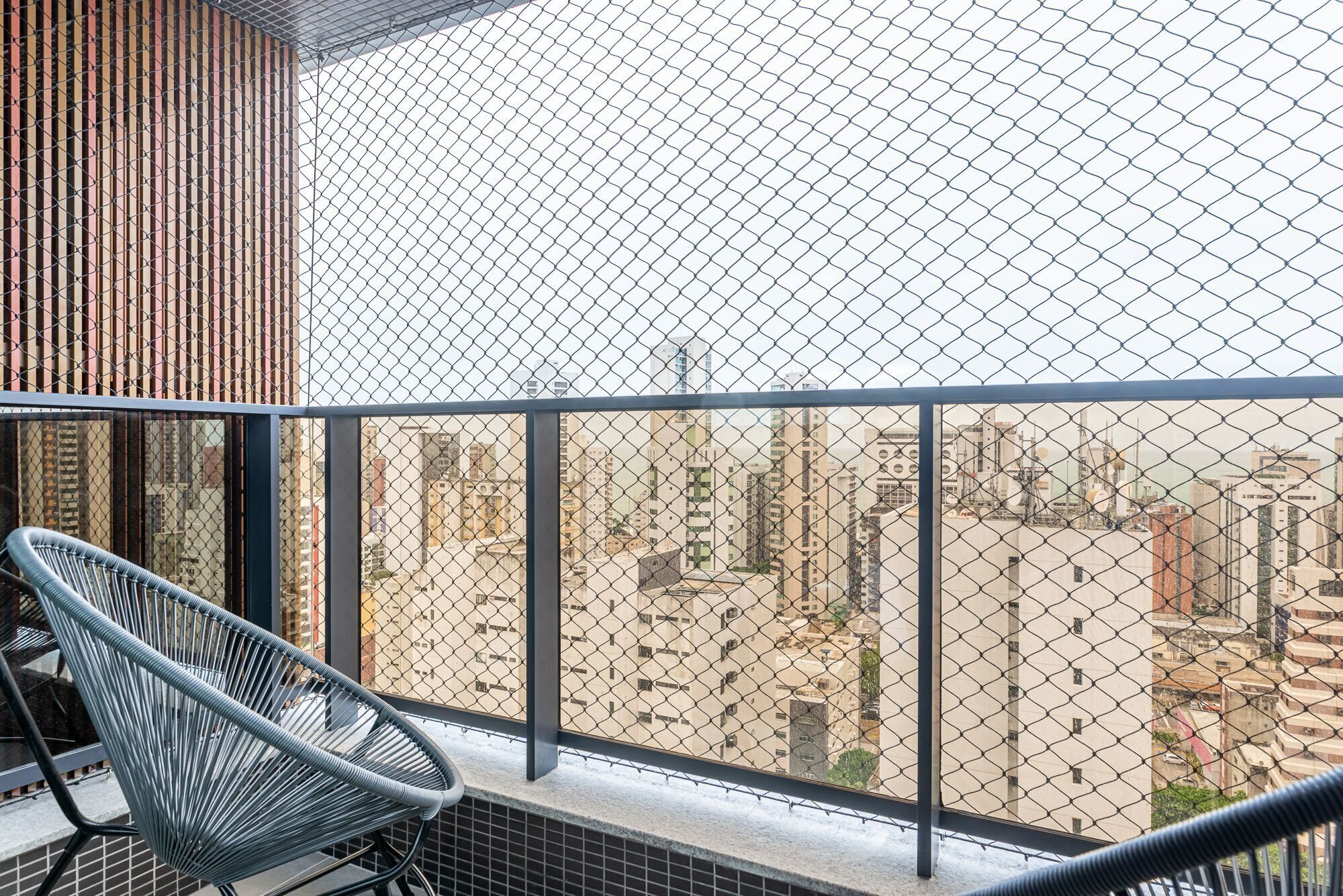 A balcony with a chain-link fence, a metal chair, and a view of a cityscape with buildings.