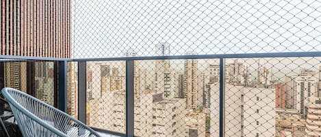 A balcony with a chain-link fence, a metal chair, and a view of a cityscape with buildings.
