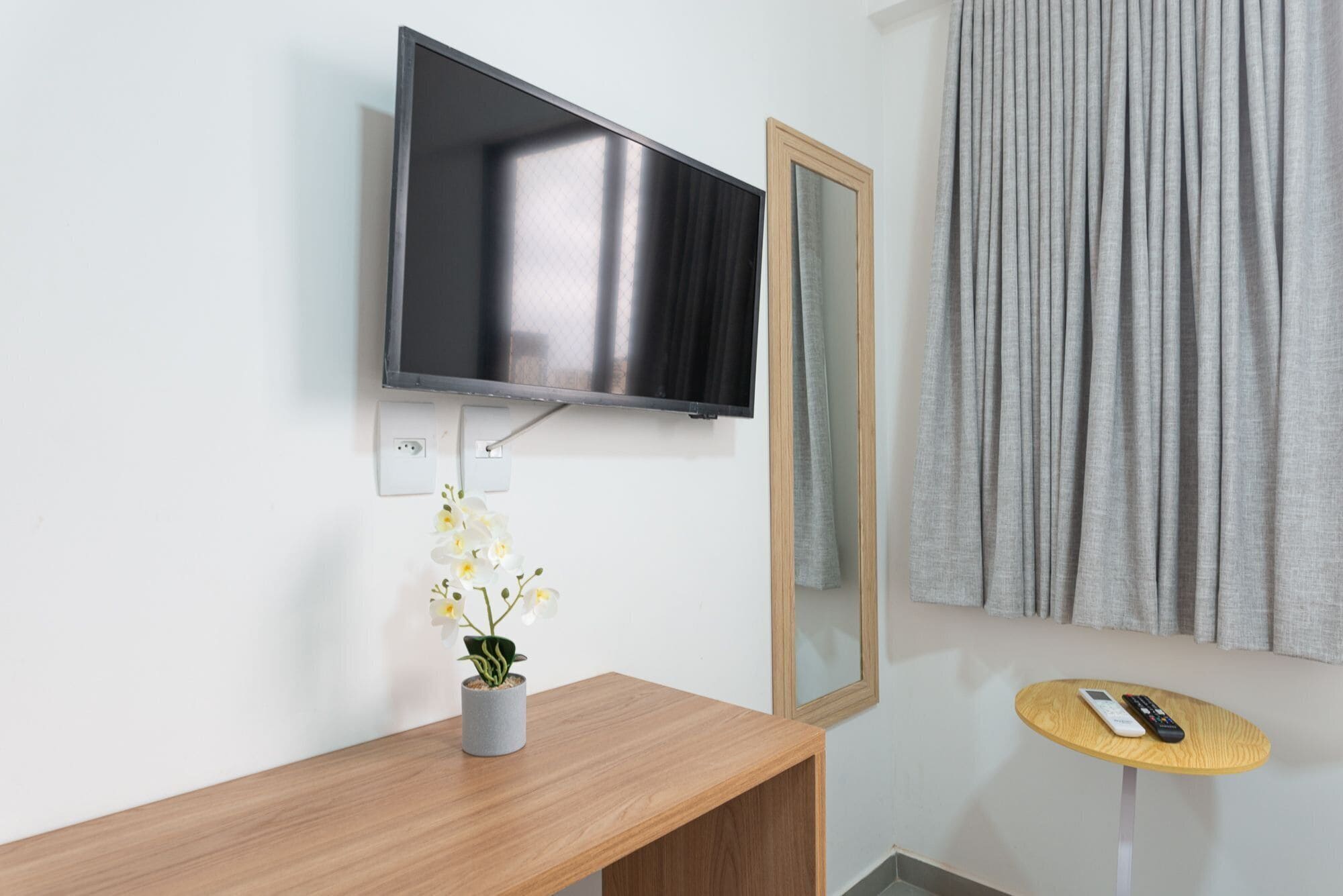 Una habitación moderna con un televisor montado, una pequeña mesa de madera, una orquídea en maceta, un espejo y una ventana con cortinas grises.