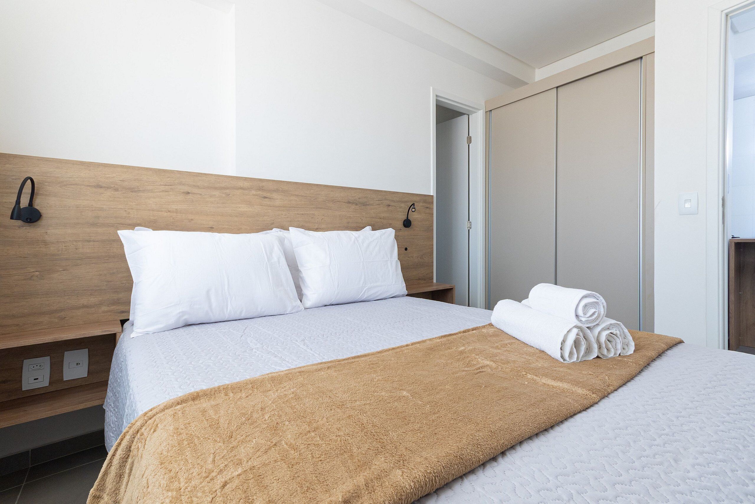 A neatly made bed with white linens, a brown throw blanket, and rolled towels on the side.