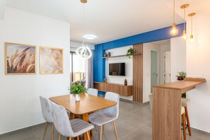 A modern kitchen with a blue accent wall, a wooden dining table with chairs, a TV mounted above the counter, and a hanging light fixture.