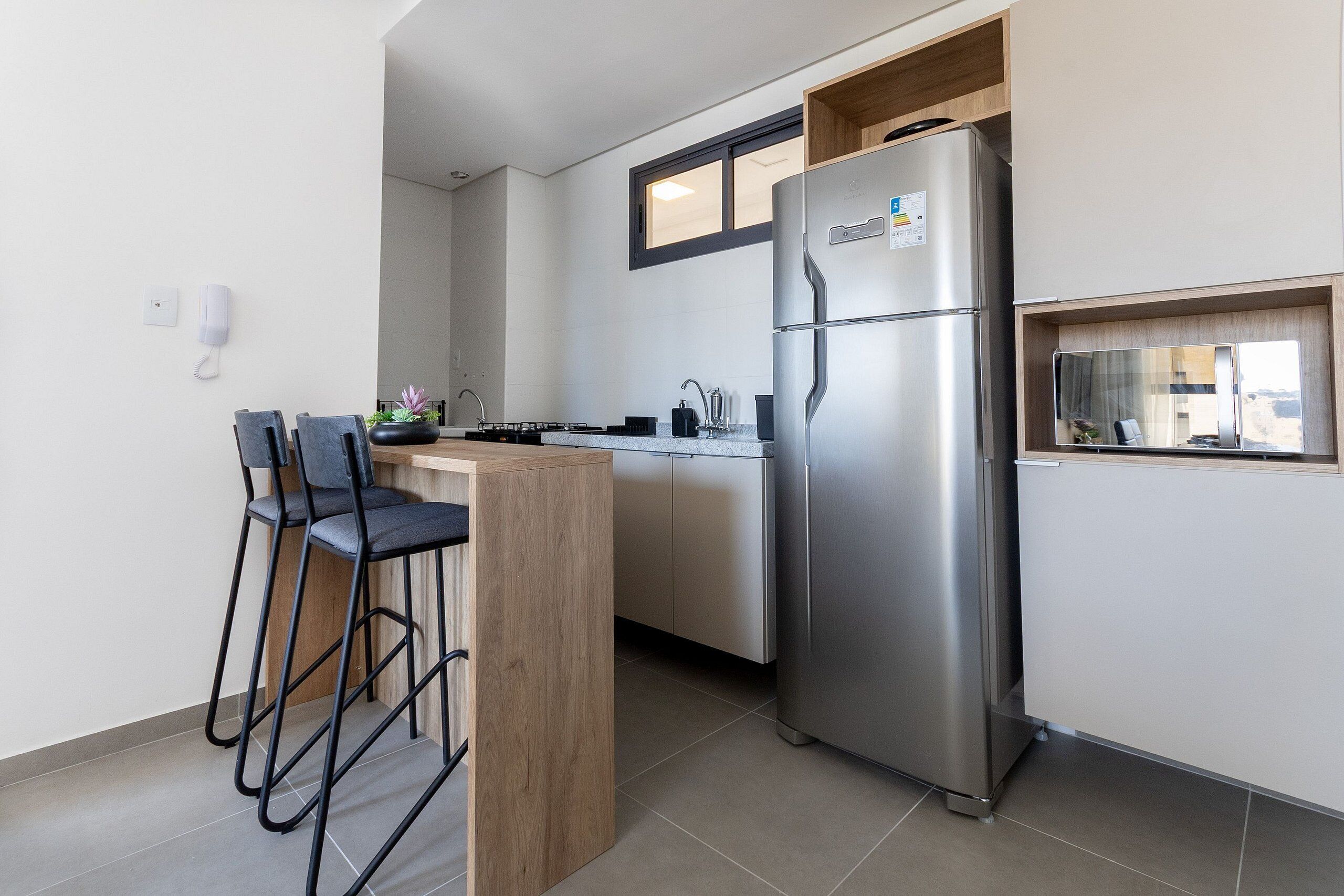 A compact kitchen with a stainless steel refrigerator, a wooden bar counter with two chairs, and a window above the sink.