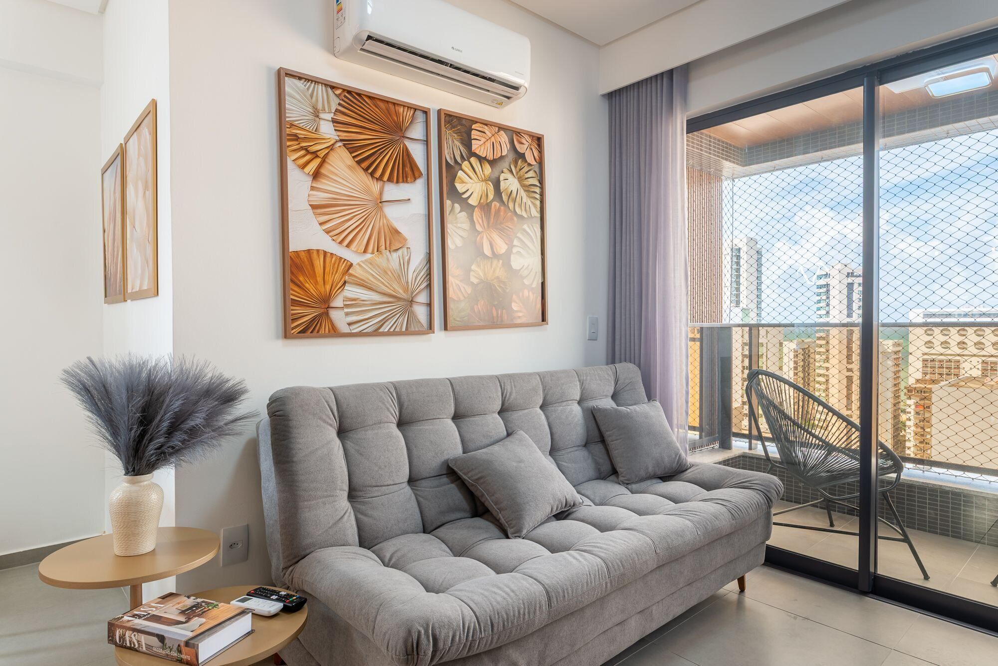 A modern living room with a grey sofa, decorative wall art, a vase with feathers, and a view of a cityscape through a balcony.