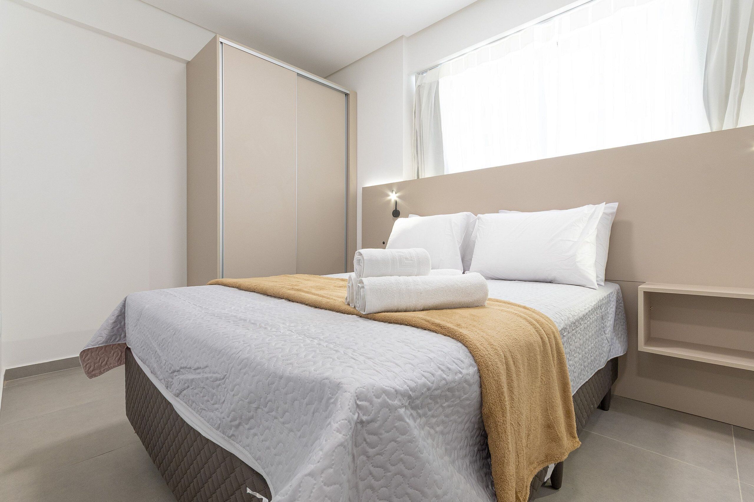 A neatly made bed with a white and beige blanket, a bedside table with a lamp, and a window with curtains.