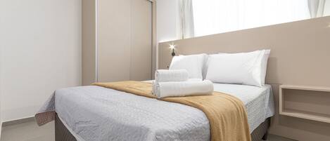 A neatly made bed with a white and beige blanket, a bedside table with a lamp, and a window with curtains.