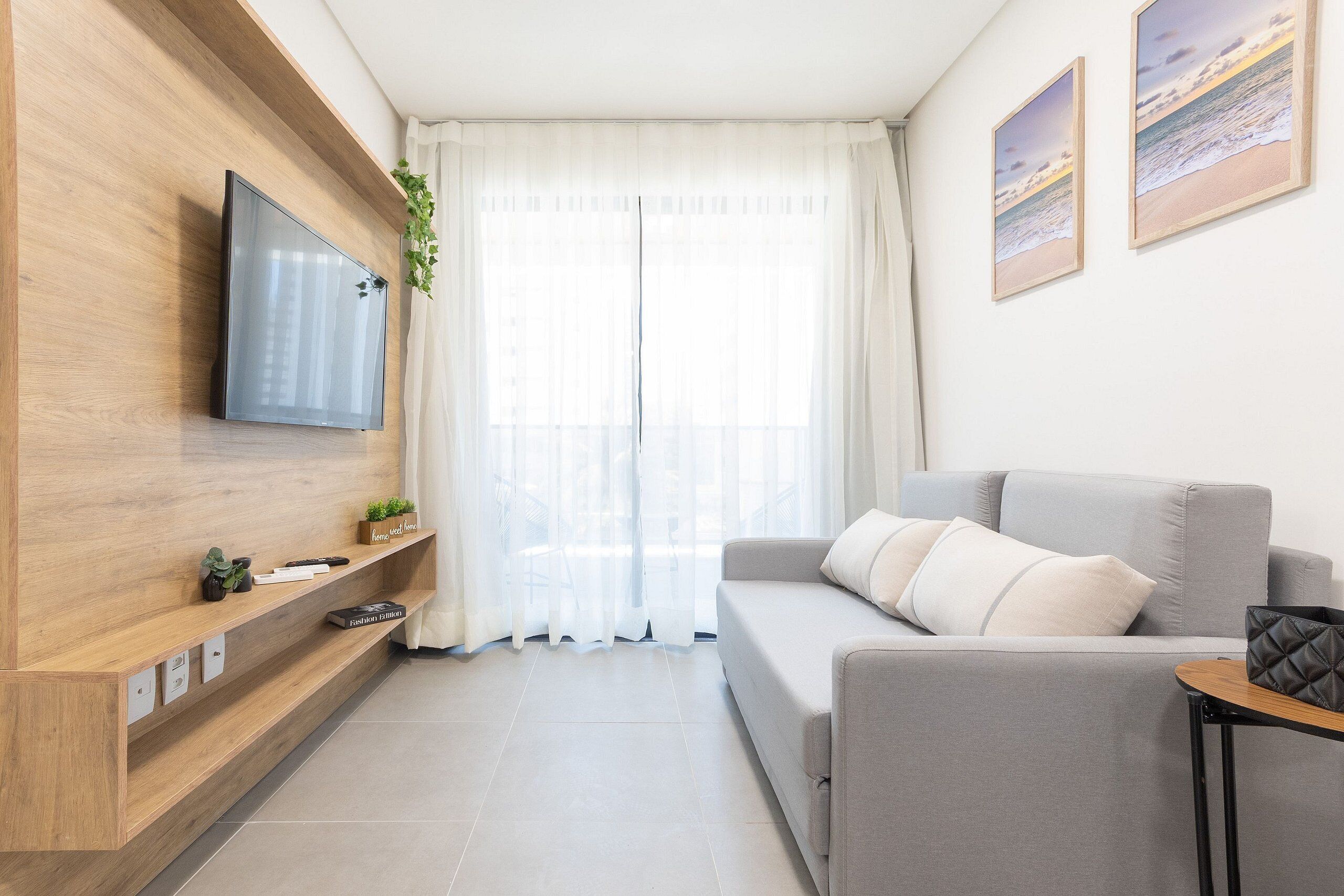 A modern living room with a grey sofa, a mounted TV, a wooden shelf with decorative items, and a large window with sheer curtains.
