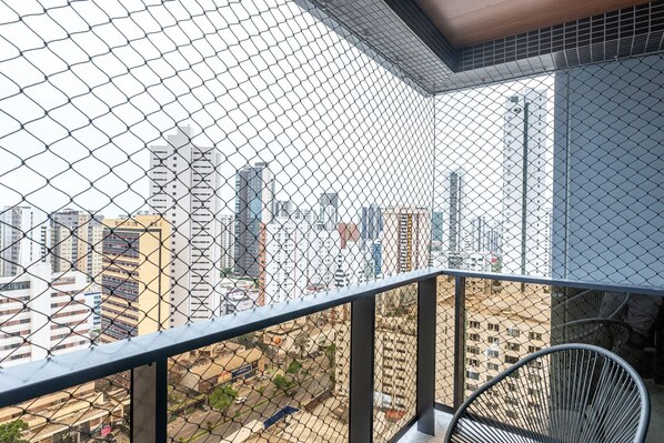 A balcony with a chain-link fence, a metal chair, and a view of a cityscape with various buildings.