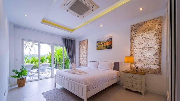 A bedroom with a bed, a nightstand, a lamp, a painting, and a view of a garden through a sliding door. - Pool Villa Black Mountain Hua Hin BM30 (Hua Hin)