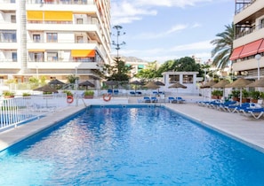 Ein Swimmingpool mit Liegestühlen und Sonnenschirmen, umgeben von einem weißen Zaun und Gebäuden mit Balkonen.