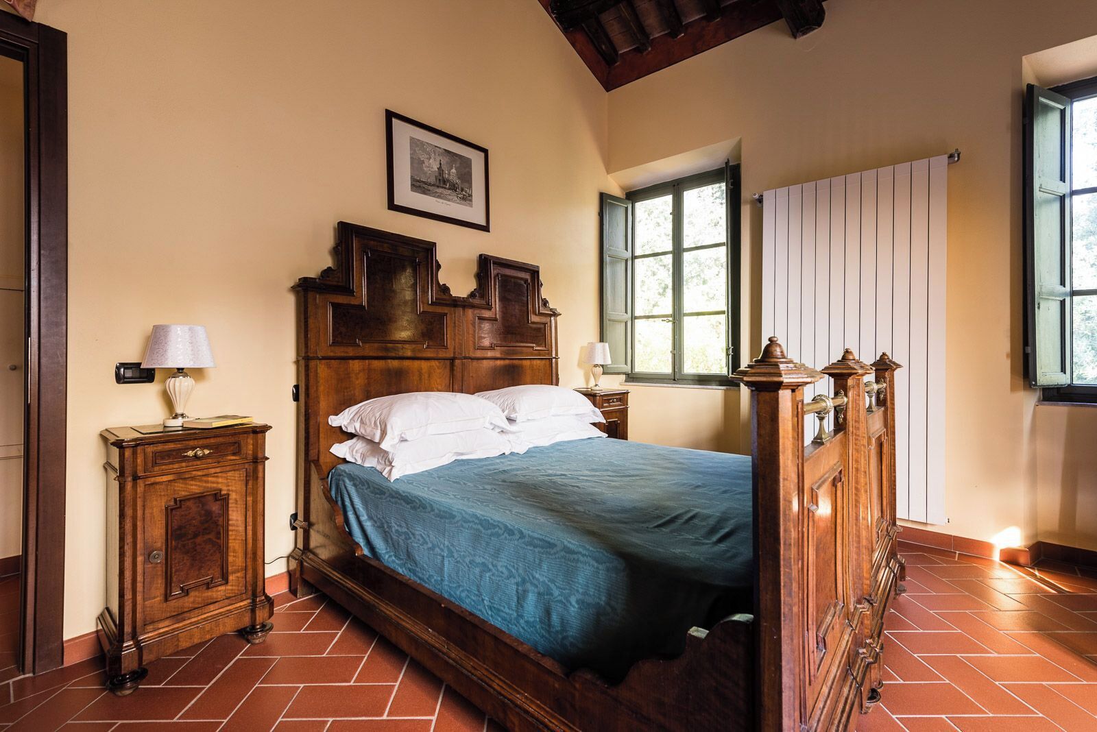 A traditional bedroom with a wooden four-poster bed, a nightstand with a lamp, a window with a view, and a radiator.