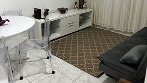 A modern living room with a white round table, clear plastic chairs, a grey sofa, and a flat-screen TV mounted on the wall. - Beachfront apartment in Gonzaguinha (São Vicente)