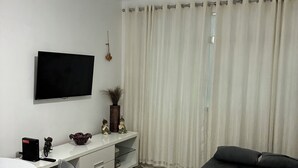 A modern bedroom with a bed, a TV mounted on the wall, a desk with a chair, and a window with curtains. - Beachfront apartment in Gonzaguinha (São Vicente)