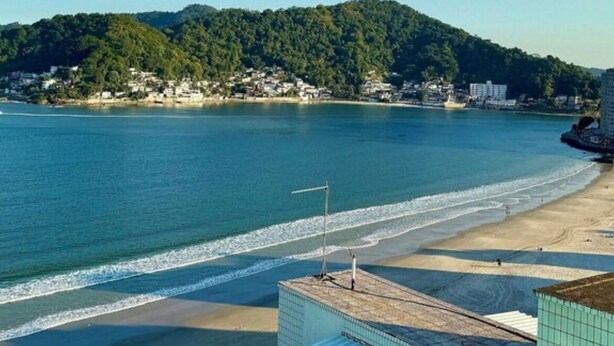 A coastal view with a beach, a clear sky, and a hill with buildings in the background. - Beachfront apartment in Gonzaguinha (São Vicente)