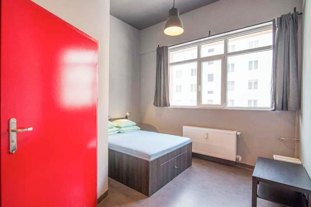 A room with a bed, a window with curtains, a radiator, and a red door.