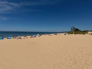 En bred, sandet strand med telte og parasoller, en klar himmel og et par både i det fjerne.