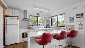 A modern kitchen with white cabinetry, a built-in oven, a stainless steel refrigerator, and a large window providing a view of the outdoors. There are three red bar stools at the kitchen island. - Family Retreat at Rhyll (Rhyll)