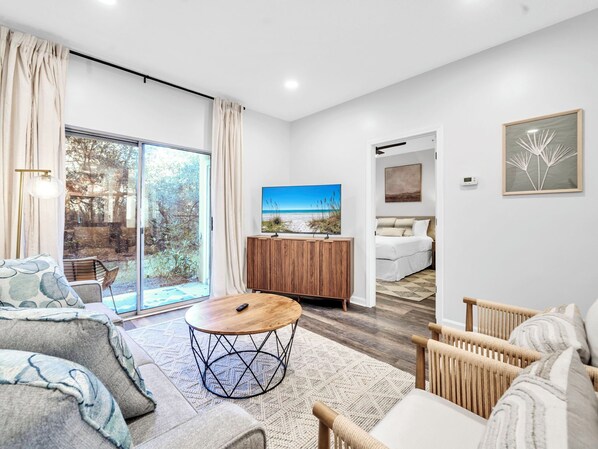 A modern living room with a large sliding glass door leading to a balcony, a wooden cabinet with a TV, and a cozy seating area with a round coffee table. - ⭐30A Heron's Watch 1102 | Sugary White Beaches⭐ (Santa Rosa Beach)