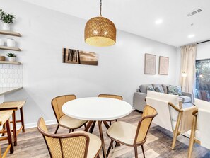 A modern dining area with a round table and wicker chairs, a hanging light fixture, and a view of the outdoors through a window. - ⭐30A Heron's Watch 1102 | Sugary White Beaches⭐ (Santa Rosa Beach)