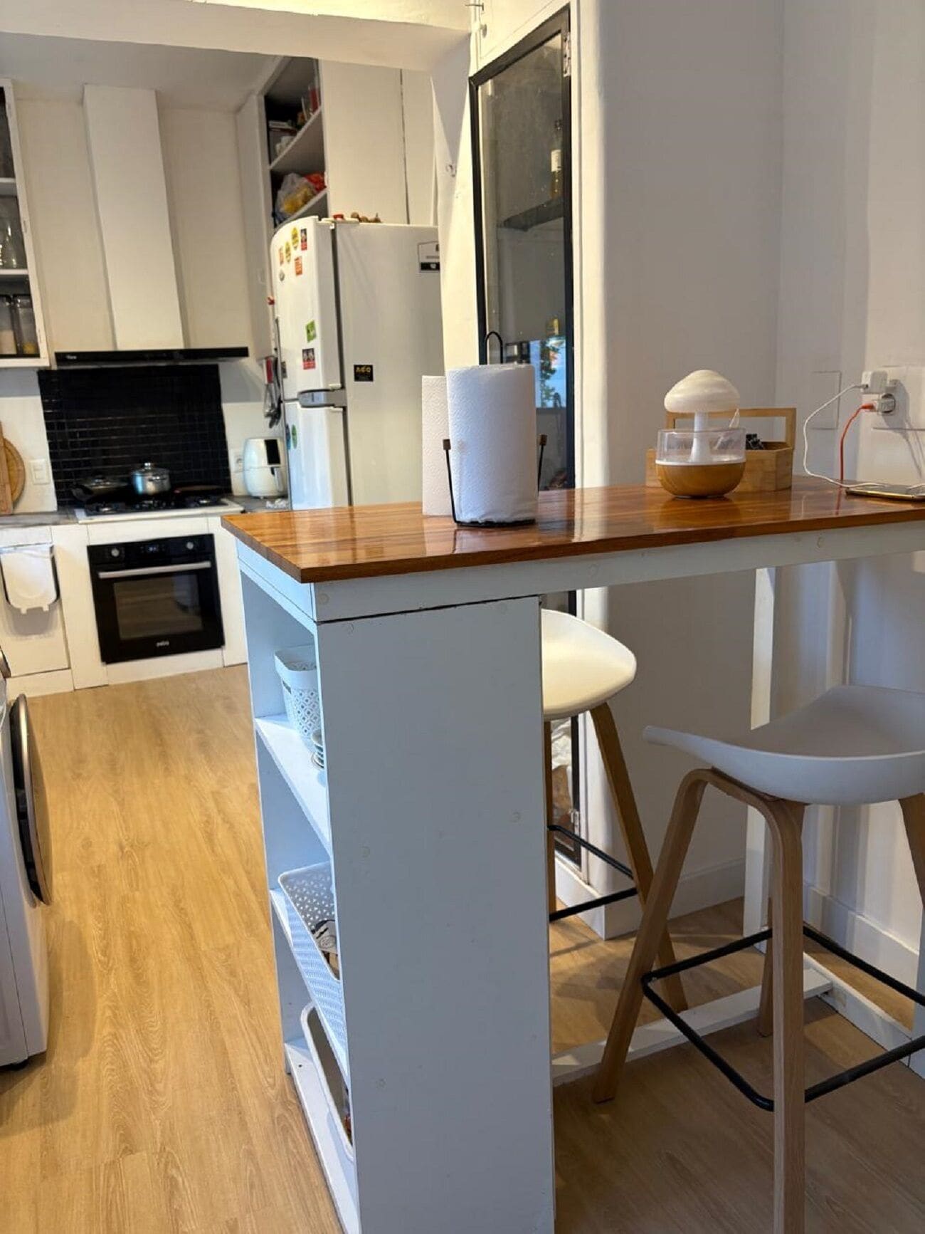 Una cocina moderna con armarios blancos, encimera de madera y horno empotrado. Hay una silla blanca y un taburete en la encimera.