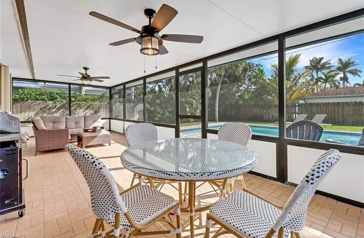 A covered patio area with a glass table and chairs, a ceiling fan, and a view of a swimming pool.