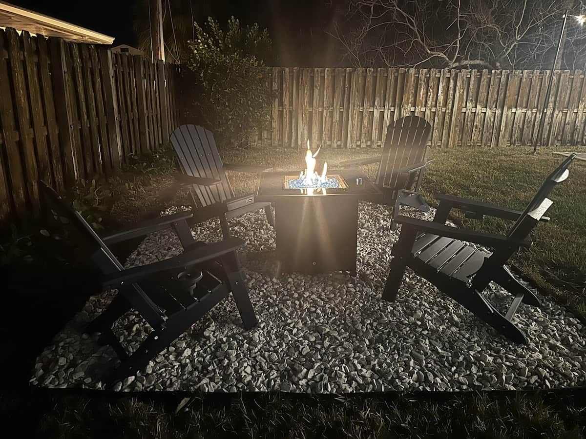 An outdoor seating area with a fire pit, surrounded by chairs and a table, set on a gravel surface.