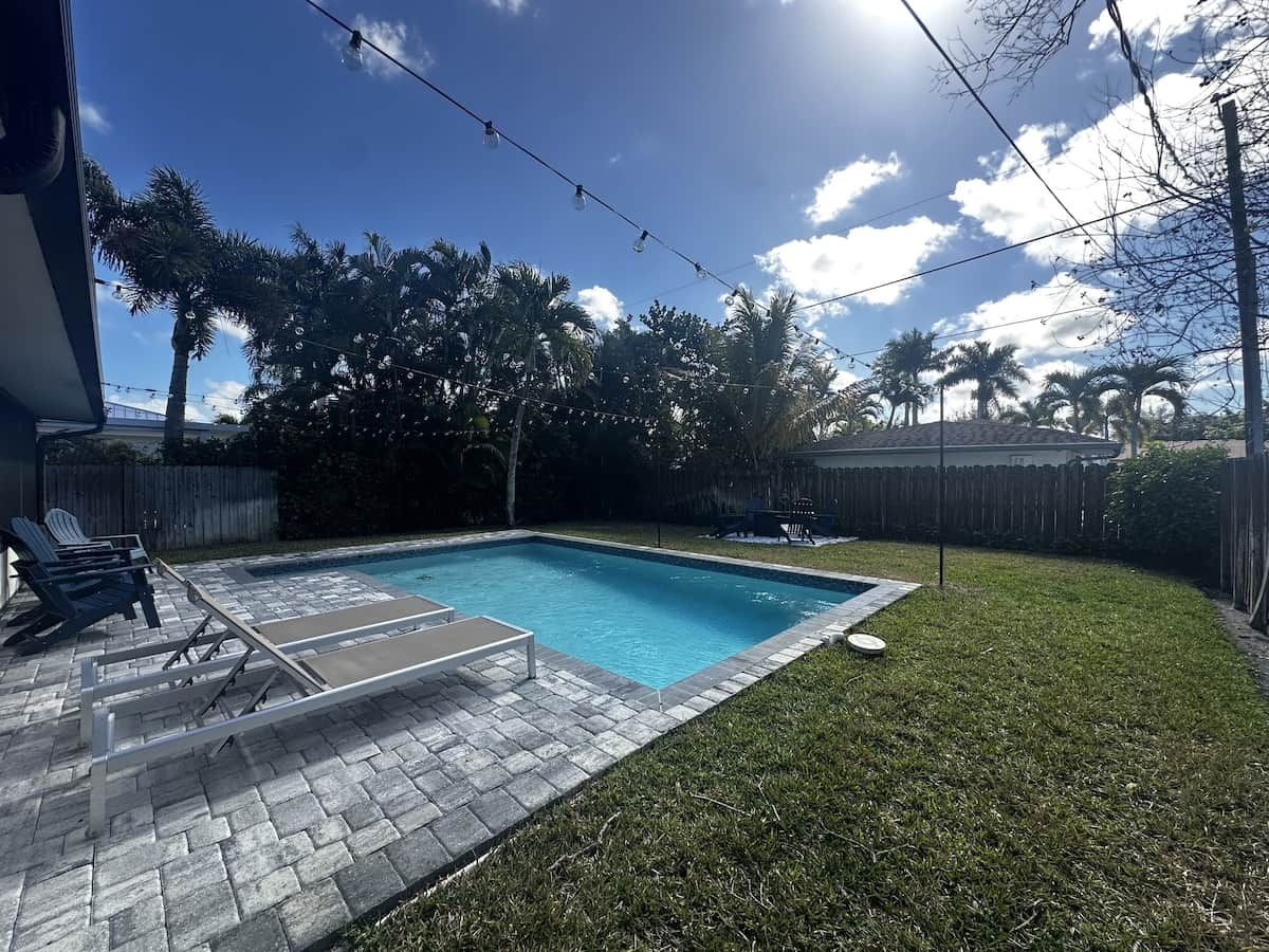 A backyard with a swimming pool, a patio with lounge chairs, and a wooden fence.