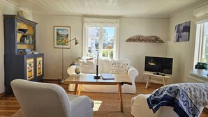 A cozy living room with a sofa, a table with a book and a candle, a TV, and a window with a view of buildings. - Charming cottage by the sea in Stocken | SE09171 (Ellös)