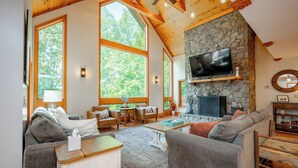 A spacious living room with a stone fireplace, large windows, and wooden ceiling beams. - Dog Friendly Lakefront Home w/ Pool & Hot Tub! (Swanton)